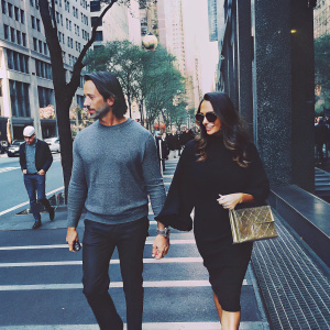Jay Rutland and Tamara Ecclestone strolling down a bustling city street. He is wearing a grey sweater and black pants. She has a black dress on and carries a gold clutch. The urban environment is filled with tall buildings and trees. People walk by in the background.