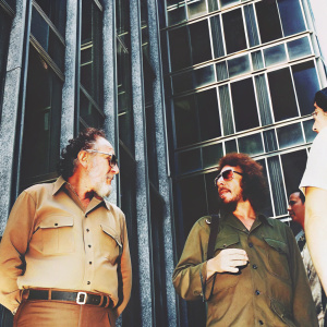 Black and white candid photo of Fidel Castro and Ernesto 'Che' Guevara standing in front of a modern skyscraper, dressed in casual attire. They appear engaged in conversation, both looking relaxed yet focused.