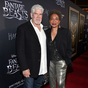 Ron Perlman and Opal Perlman at a Hollywood event. Ron in a white shirt and dark blazer, Opal in a black jacket with metallic sequins and silver pants. 'Fantastic Beasts' promotional backdrop