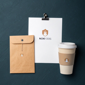 Promotional setup for Noki Dog. Includes brown envelope, white paper, small business card, and coffee cup, all branded with Noki Dog logo. Dark background with subtle texture.