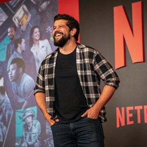 A casual shot of a Hispanic man with a full beard. Wearing a black t-shirt with a graphic design and a plaid shirt over it. Dark jeans and a big smile. The background is filled with the vibrant Netflix logo and imagery.