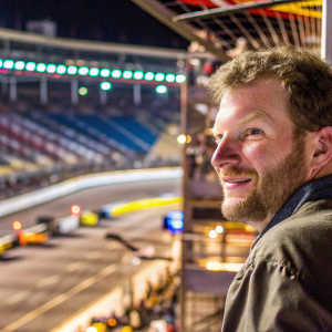 Dale Earnhardt Jr., with an engaging smile, captures the essence of motorsport enthusiasm. Posed casually with blurred stadium background.