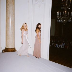 Victoria Silvstedt and Rachel Legrein Trapani at a gala event. Intricate chandeliers and grand columns. Victoria with flowing blonde curls, Rachel with sash, both posing elegantly. Crowded room with onlookers.