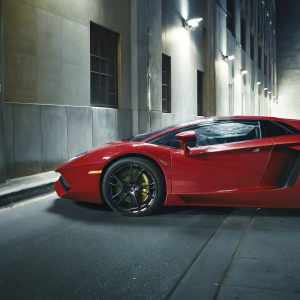 A sleek red Lamborghini Aventador LP700-4 parked on a grey urban street. Black and gold accents contrast against the red car body. Neutral color scheme.