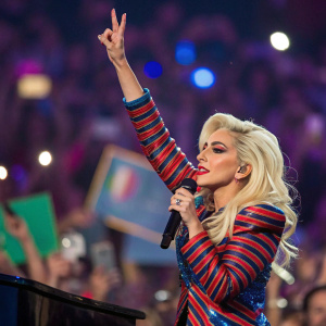 Lady Gaga, blonde, striking a peace sign pose. Onstage during concert, crowd visible