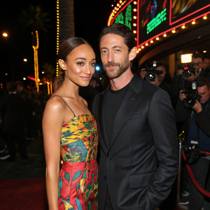 Iddo Goldberg and Ashley Madekwe at the Hollywood party. Ashley in a colorful, vibrant dress. Iddo in a sleek black suit. Neon lights and paparazzi in background.