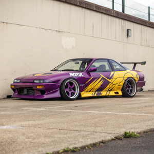 A Nissan 180SX Rocket Bunny, decorated with Motul logo, parked against a plain wall. Bright purple and yellow paint jobs stand out, making the car look like a character from a fantasy world.