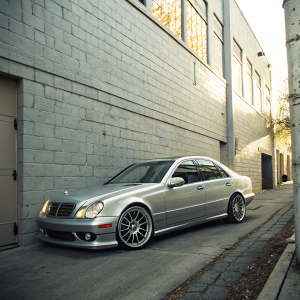 A silver 2003 Mercedes E55 AMG parked in a quiet, sunlit alley. Gray brick walls provide a serene backdrop. Car's sleek chrome rims and accents glisten under the natural light, casting soft shadows that accentuate its contours.