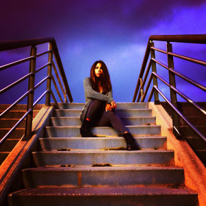Meaghan Rath as Sally Malik from 'Being Human', sitting on a concrete staircase. Dark, dramatic setting with metal railings. She's wearing a gray sweater and leggings. Serious expression. Background blurred.