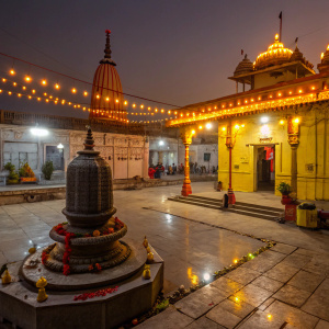 Maha Shivratri festival scene. Nighttime, temple courtyard. Luminous yellow and orange lights. Shiva lingam and statues. Traditional offerings and decorations.