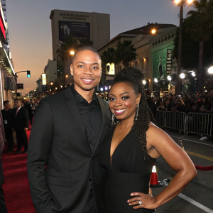 Ian Wallace and Jemele Hill at a Hollywood premier. Wearing black dress and suit. Background is crowded street.