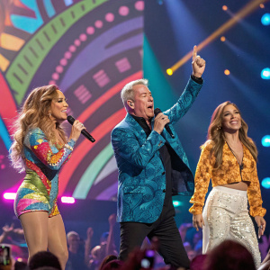 Jennifer Lopez, Elvis Duran, and Carolina Bermudez on the lively iHeartRadio stage. Bright lights and audience in the background.