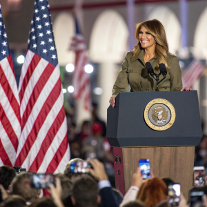 Melania Trump, smiling, standing at podium with American flags on either side. Crowded event, people taking photos