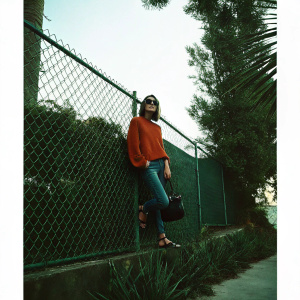 Cobie Smulders, casually dressed in an orange sweater and sunglasses, leans against a chain-link fence. Jeans, sandals, and black handbag complete her look in a candid photo.