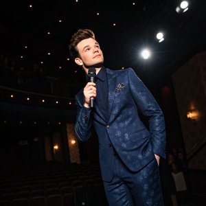 Chris Colfer, at the Gotham Awards, wearing a navy blue suit with a subtle pattern. Poses elegantly with a microphone in hand. Dark room with spotlights, no audience visible