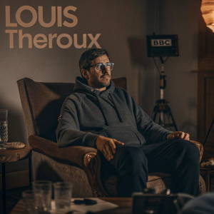 Louis Theroux, seated in a cozy living room, donning a casual grey hoodie and glasses. Warm lighting and a light-colored wall backdrop. The setting exudes a friendly, intimate vibe. BBC logo and show name prominently displayed.