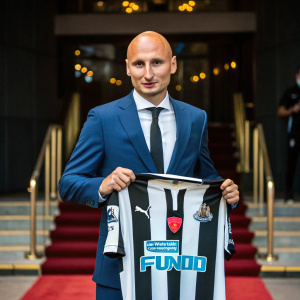 Jonjo Shelvey, bald head, wearing a blue suit. Holding the Newcastle United jersey. Formal event backdrop
