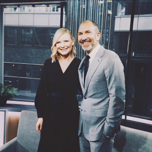 Kirsten Dunst in a black dress with Matt Lauer on a talk show set. Friendly expressions, warm smiles. Modern studio setting with soft lighting and building-like backdrop.