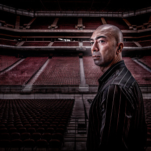 Roberto Di Matteo, former Chelsea FC manager, bald head, intense gaze. Dressed in a black and white striped shirt. Standing alone in an empty stadium.