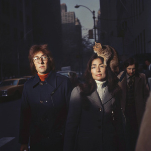 John Lennon and Yoko Ono walking down a bustling New York City street. A cat perches on John's shoulder, amidst the urban backdrop.