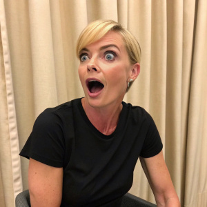 Close-up shot of Jaime Pressly. She has a surprised expression. Wearing a black t-shirt and sitting in front of a beige curtain.
