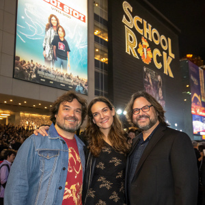 Jack Black, Rebecca Brown, and Joey Gaydos at the School of Rock premiere. They are in casual outfits, posing in front of a large movie poster. Brightly lit, crowded event space in Tokyo.