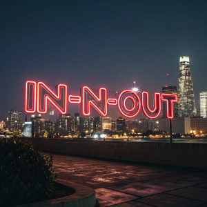 In-N-Out Burger logo, featuring the brand name 'In-N-Out' in a vibrant neon font, set against a dark urban night skyline.