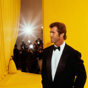 Mel Gibson at the Academy Awards, dressed in a tuxedo. The red carpet is bustling with photographers.