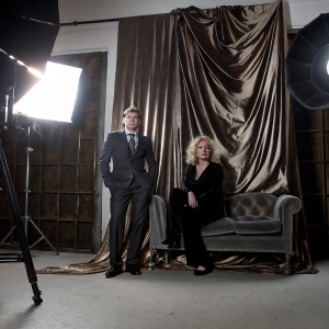 Barry Pepper and Cindy in a glamorous studio setting. Dark suit, tie, styled haircut. Cindy's curly blonde hair, tailored dark outfit. Rich red velvet background