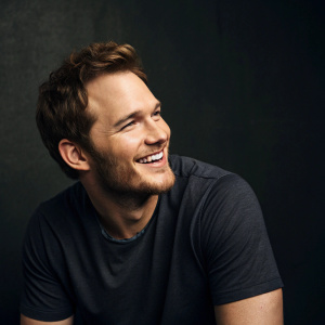 Chris Pratt, actor, in a candid photo. Poses with a warm smile. Dark studio backdrop, minimalistic setting.