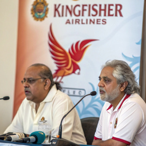 Vijay Mallya and Captain Gopinath at a press conference. They sit at a table with microphones in front of them. Behind them, a large banner featuring Kingfisher Airlines logo.