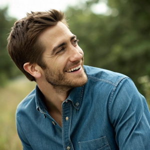 Jake Gyllenhaal, with a charming smile, wearing a casual blue denim shirt. Set against a blurred greenery backdrop.