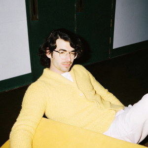 Joe Jonas in a casual setting. Wearing a green knitted sweater and glasses. Simple, light background