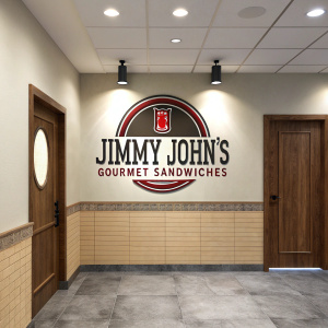 Stylized Jimmy John's Gourmet Sandwiches logo with dark monogram in circular design, accompanied by text. Featured in a cozy, contemporary office space. Neutral colored walls, wooden doorway, and tiled accents. Spotlit center piece