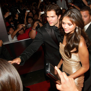 Nina Dobrev and Henry Cavill attending a bustling film festival. Both are seen mingling with fans and paparazzi. Nina is dressed in a shimmering gold gown, while Henry is in a tailored black tuxedo.
