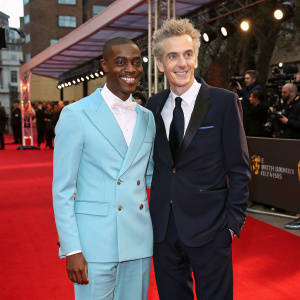 Ncuti Gatwa and Peter Capaldi at the BAFTA awards. Ncuti in a light blue suit, Peter in a dark suit. They are standing on a red carpet with flashing cameras in the background.