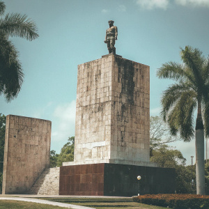 Che Guevara's mausoleum in Cuba. A massive stone monument with a statue depicting a determined figure standing atop a rock. The backdrop features a clear blue sky, lush greenery, and towering trees. The structure is part of a sprawling complex set within a serene park.