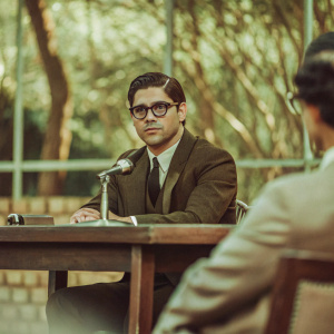 Wilmer Valderrama at a Q&A session. Wearing a dark suit and glasses. Serious expression.