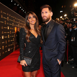 Antonella Roccuzzo, in a black sequined dress and clutch, alongside Lionel Messi in a suit. They pose confidently on a bustling red carpet. Dimly lit background with various logos and camera flashes.