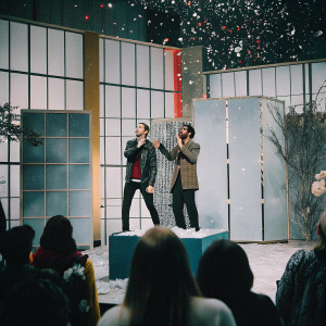 Rhett James McLaughlin and Charles Lincoln Neal III, better known as Rhett & Link, are hosting an energetic talk show. A bright, colorful set with a lively audience.