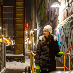 Anna Kendrick standing in a gritty urban setting. Graffiti-covered walls, serious expression. White beanie, black jacket with fur trim, green gloves.