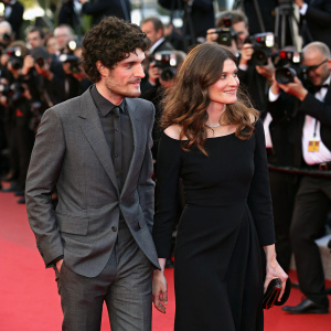 Louis Garrel and Chiara Mastroianni at Cannes Film Festival premiere. Louis in grey suit, Chiara in black dress. Red carpet and flashing cameras.