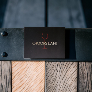 Business card for 'Choors Lah!', sleek design with wooden texture background. Red wine symbol and text in earthy tones. Photo-style graphic