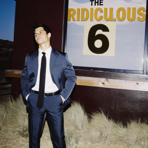 Taylor Lautner in dark blue suit, white shirt, and black tie, standing confidently in front of 'The Ridiculous 6' movie poster. Rustic film-themed backdrop