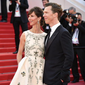 Benedict Cumberbatch and Sophie Hunter at the Cannes Film Festival. Benedict in a sleek black suit. Sophie in an elegant white dress with intricate floral designs. Background features bustling red carpet and flashing cameras.