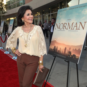 Maria Conchita Alonso at the movie premiere of 'Norman'. Wearing a cream blouse with lace overlay and brown pants. She sports glittery accessories and smiles warmly. The movie poster is displayed on an easel-like stand. The scene is set on a red carpet with a glamorous atmosphere.