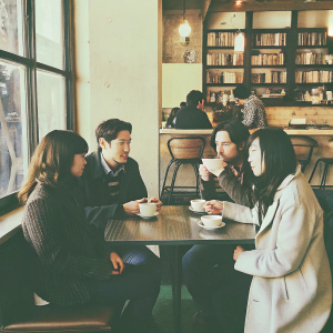 At a bustling cafe, actors Teo Yoo, Greta Lee, John Magaro, and Celine Song gather for an informal meet-up. They are seated at a cozy table with cups of coffee.