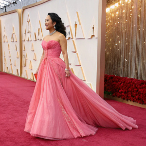 Stephanie Hsu on the Oscars red carpet. Wearing a flowing, strapless, pink gown. Backdrop of shimmering golds and warm hues.