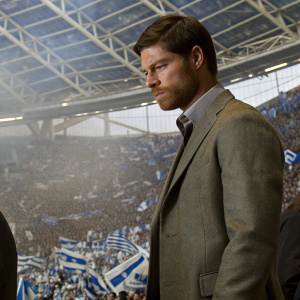 Xabi Alonso, looking serious, dressed in a grey suit. Inside an arena, amidst cheering fans.