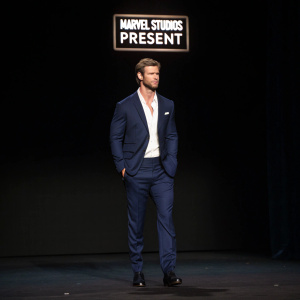 Chris Hemsworth in a navy suit with a white shirt. Standing alone on a stage. Behind him is a stark black background with glowing white text that reads 'Marvel Studios Present'.